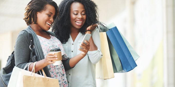 two woman smiling looking down at one of their smartphone's while holding shopping bags and a hot beverage